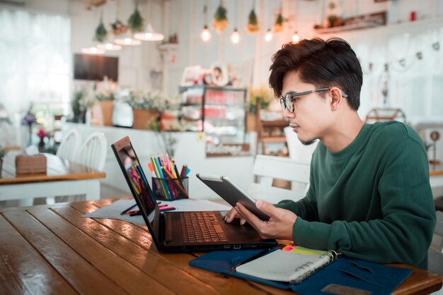 Azjatycki męski uczeń pracuje używać laptopu obsiadanie przy drewnianym stołem w sklep z kawą