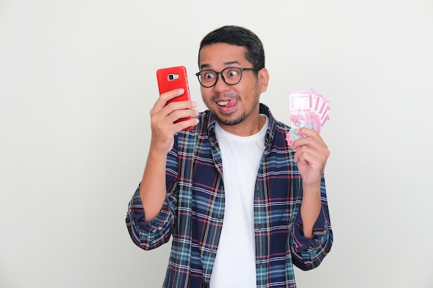 Azjatycki mężczyzna patrzy na swój telefon z zabawnym wyrazem twarzy, trzymając pieniądze