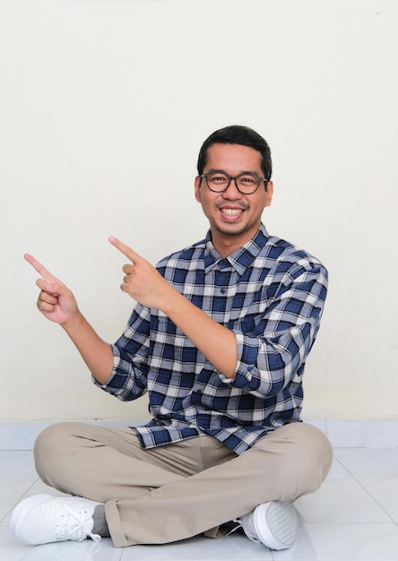 Azjatycki mężczyzna siedzi na podłodze, wskazując prawą stronę z radosnym wyrazem twarzy