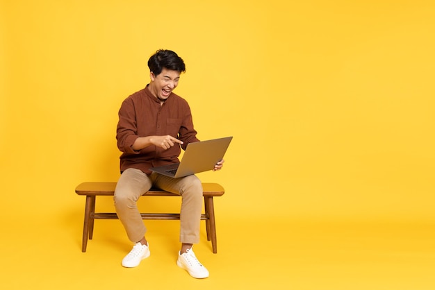 Azjatycki mężczyzna trzyma laptopa i siedzi na drewnianym krześle na białym tle na żółtym tle