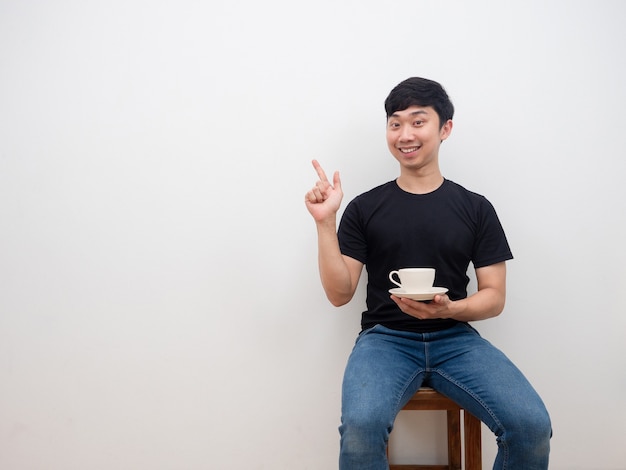Azjatycki mężczyzna trzymający filiżankę kawy i wskazujący palcem w przestrzeń ze szczęśliwą uśmiechniętą twarzą siedzącą na krześle na białym tle