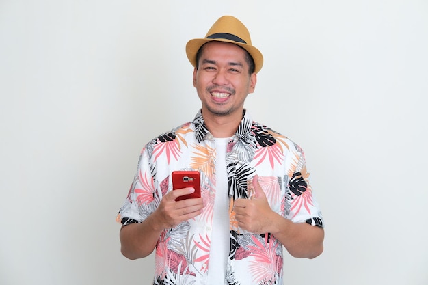 Azjatycki mężczyzna ubrany w plażową koszulę i kapelusz daje kciuk do góry, trzymając swój telefon