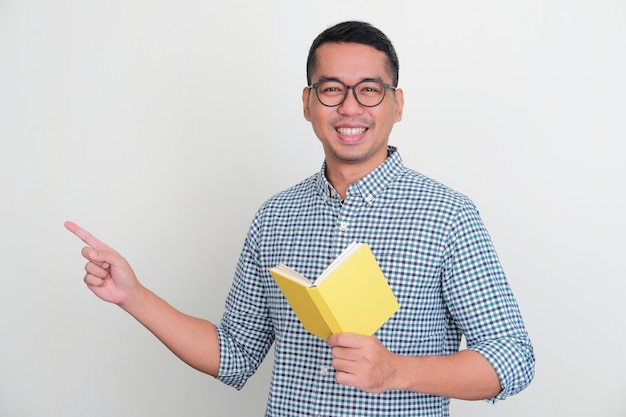 Azjatycki mężczyzna uśmiechający się i wskazujący na prawą stronę, trzymający książkę