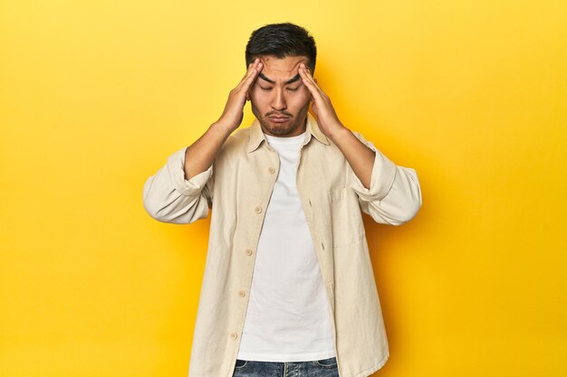 Azjatycki mężczyzna w otwartej koszuli, białej koszulce na żółtym studio dotyka świątyń i ma ból głowy.