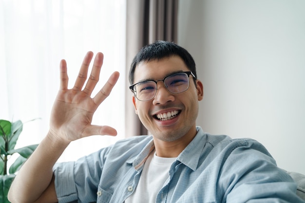 Azjatycki mężczyzna za pomocą smartfona do wideokonferencji z przyjaciółmi macha ręką, wykonując gest powitania