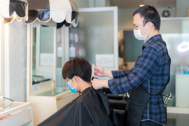 Azjatycki młody mężczyzna i fryzjer noszący maskę medyczną w celu ochrony podczas powieści Coronavirus, Covid-19 w Barbershop Hair Care Service.