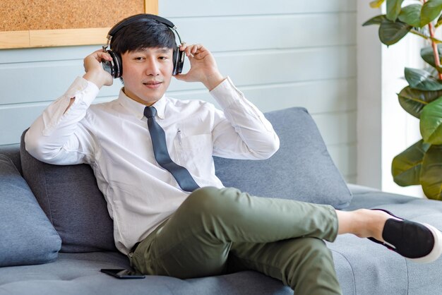 Azjatycki młody przystojny zawodowy udany mężczyzna biznesmen pracownik w formalnej koszuli biznesowej i krawat, siedząc na wygodnej kanapie, słuchając strumieniowego przesyłania muzyki online przeglądanie Internetu za pomocą smartfona.