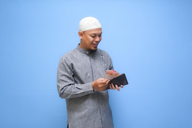 Zdjęcie azjatycki muzułmanin trzymający portfel pełen gotówki gotowy do zapłaty zakatu wyizolowany na niebieskim tle