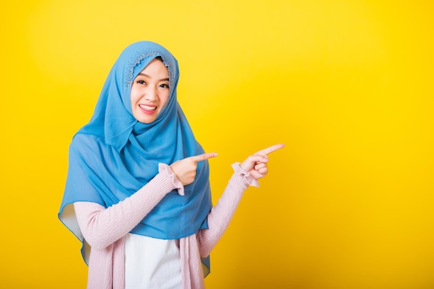 Azjatycki muzułmański Arab, Portret szczęśliwej pięknej młodej kobiety Islam religijny nosić welon hidżab zabawny uśmiech ona pozytywny wyraz wskazujący palcem bokiem do przestrzeni na białym tle żółte tło