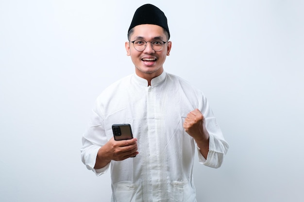 Azjatycki muzułmański mężczyzna trzymający smartfona robi zwycięski gest trzymający telefon komórkowy Szczęśliwy otrzymaj specjalny prezent online na białym tle