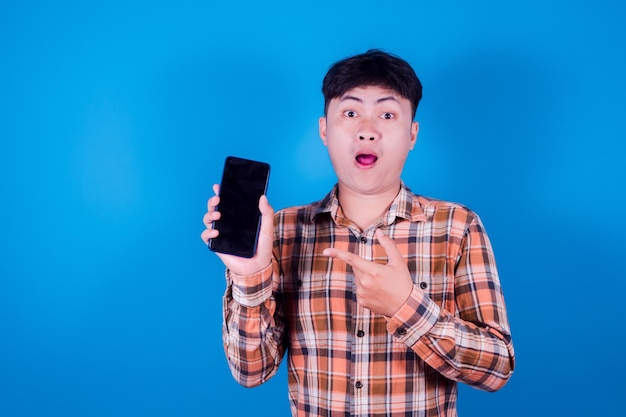 Azjatycki portret młodego białego człowieka stojącego nosić koszulę, co palcem wskazującym na inteligentny cyfrowy telefon komórkowy pusty ekran na niebieskim tle