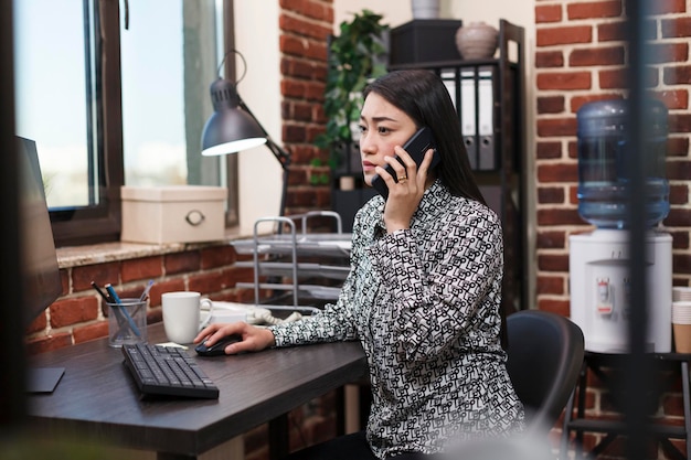 Azjatycki pracownik biurowy rozmawia przez telefon z liderem zespołu w miejscu pracy o niedokończonej prezentacji projektu. Młoda businesswoman za pomocą telefonu komórkowego, aby poinformować zespół marketingowy o złym raporcie strategii.