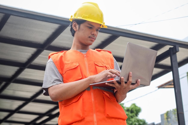 Azjatycki pracownik budowlany noszący żółty kask dla bezpieczeństwa i korzystający z laptopa na placu budowy