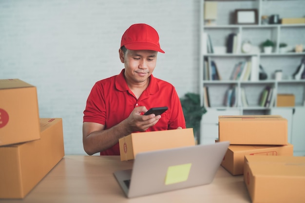 Azjatycki pracownik dostawy uśmiechający się za pomocą mobilnego skanowania kodów kreskowych dla pudełek do wysyłania lub przenoszenia paczek pocztą Noszenie czerwonego munduru praca w domu