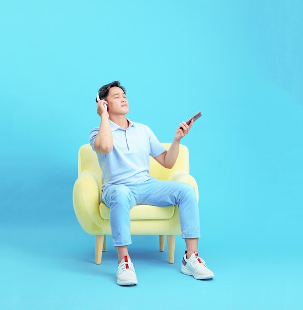 Azjatycki przystojny i wesoły młody mężczyzna siedzący na relaksującej kanapie używa smartfona i odtwarza muzykę na białym tle