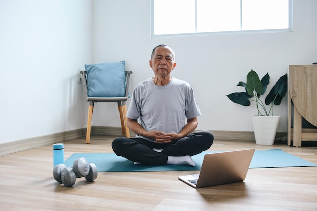 Azjatycki starszy mężczyzna medytujący na matce uczy się jogi online używając laptopa w domu