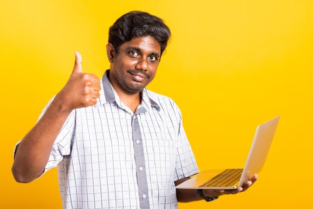 Azjatycki szczęśliwy portret młodego czarnego mężczyzny uśmiechnięty stojący nosić koszulę za pomocą laptopa i pokazujący kciuk do góry patrzący na kamerę na białym tle, studio strzał żółte tło z miejsca na kopię