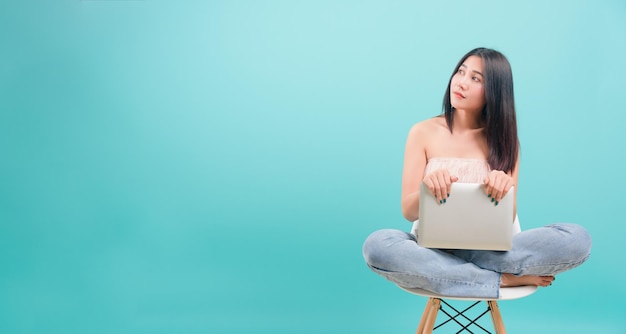 Azjatycki szczęśliwy portret piękna młoda kobieta siedząca na krześle uśmiecha się, trzymając laptopa i patrząc na bok na niebieskim tle z miejsca kopiowania tekstu