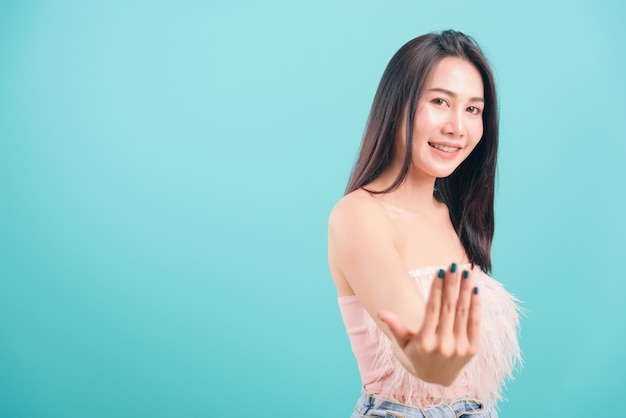 Azjatycki szczęśliwy portret piękna młoda kobieta stojąca uśmiechnięty gest pozytywny ręką zapraszającą do przyjścia tutaj znak i patrząca w kamerę na niebieskim tle z miejsca kopiowania tekstu