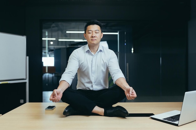 Azjatycki szef biznesmen medytujący w biurze mężczyzna siedzący w pozycji lotosu na biurku
