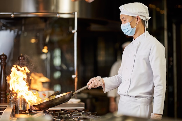 Azjatycki szef kuchni w masce oddechowej robi stirfry w płonącej patelni