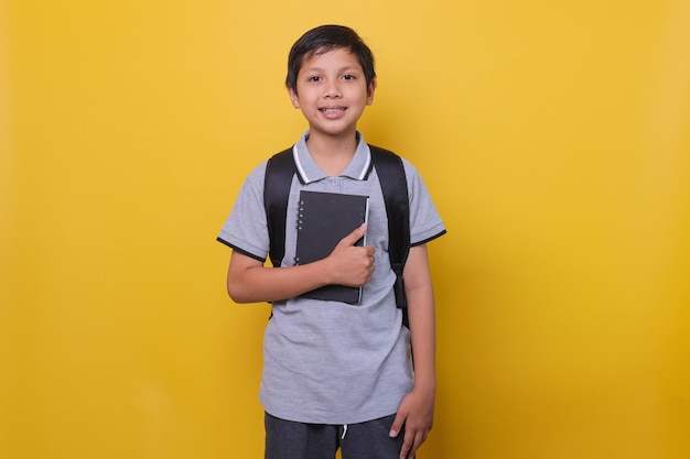 Azjatycki szkolny chłopiec w swobodnym stylu z czarnym plecakiem uśmiecha się i trzyma książkę odizolowaną na krzyczeć