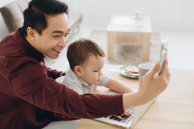 Azjatycki tata i jego synek ma śniadanie przy laptopie i bierze selfie na telefonie