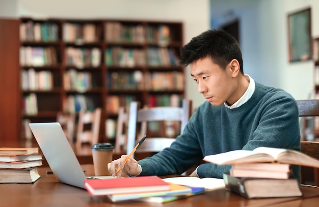 Azjatycki uczeń studiuje w bibliotece z laptopem