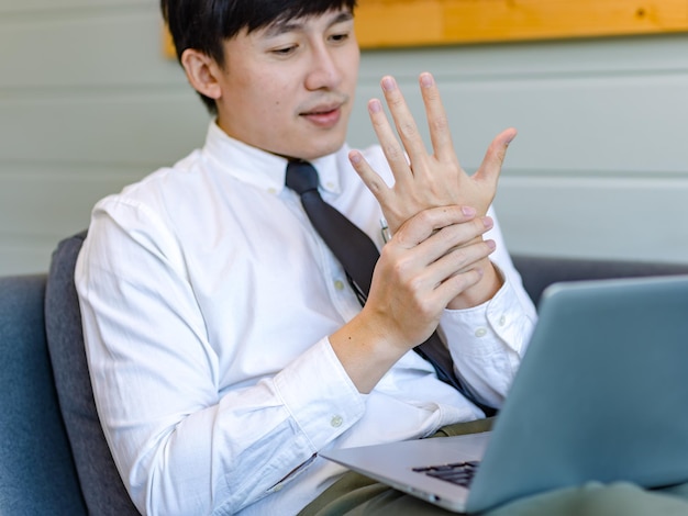 Azjatycki zmęczony przepracowanie mężczyzna biznesmen pracownik w stroju biznesowym siedzi na kanapie pracując z laptopem trzymając dłoń masaż na sztywny uraz szyi i barku z zespołu biurowego.