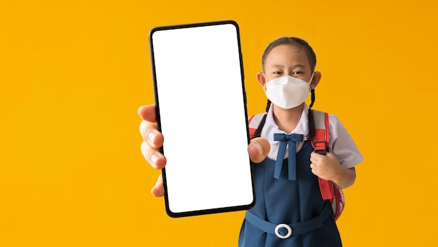 Azjatyckie dziecko uczennica uczennica nosi mundur w medycznej masce ochronnej, trzymając duży smartfon na żółtym tle na białym tle Ścieżki przycinające do prac projektowych pusta wolna przestrzeń makieta wyświetlania produktu