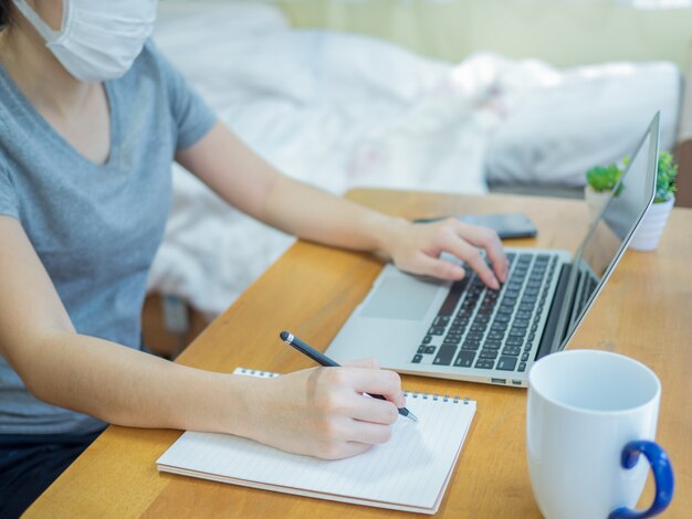Azjatyckie kobiety noszące maski pracują w domu z laptopem, pisząc w notatniku, aby ograniczyć rozprzestrzenianie się koronawirusa