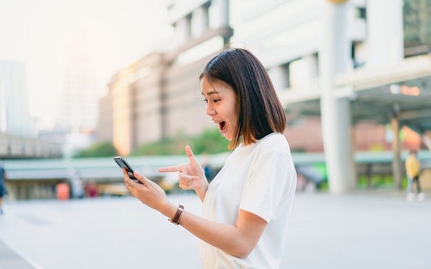 Azjatyckie kobiety szczęśliwy ono uśmiecha się i trzyma mądrze telefon z zadziwiający dla sukcesu.