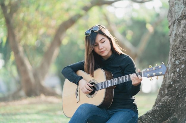 Azjatyckie muzyczki Ubrane w czarną koszulę z długimi rękawami i okulary przeciwsłoneczne na głowie Śpiewają i grają na gitarze akustycznej pod drzewem w parku rano