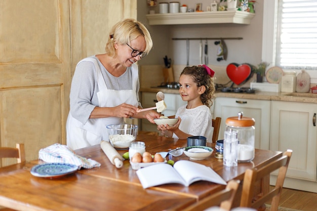 Babcia i śliczna kędzierzawa wnuczka przygotowują razem w kuchni, ze składnikami na stole.