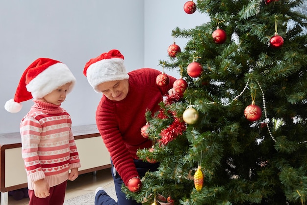 Babcia i wnuczka w czapkach Mikołaja i czerwonych swetrach ubierają choinkę w świąteczne zabawki