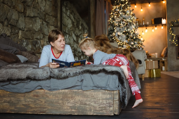 Zdjęcie babcia i wnuczka w świątecznej piżamie czytają książkę, leżąc na łóżku w domku. rodzinna koncepcja bożego narodzenia.