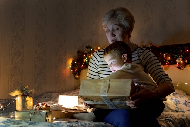 Babcia i wnuk siedzą na sofie otwierając świąteczny prezent w pokoju z jasnymi girlandami