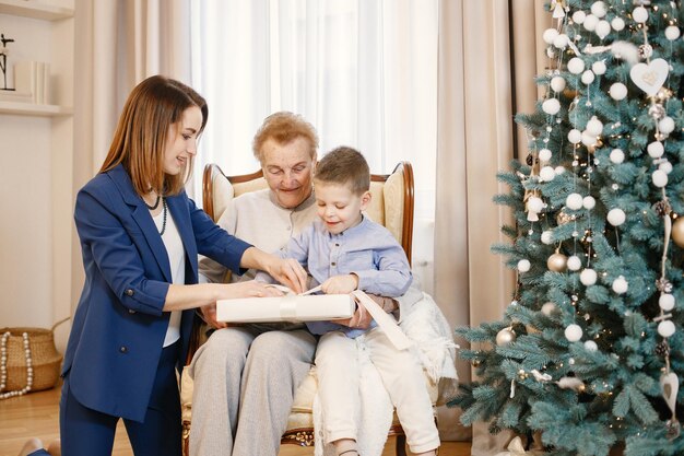 Zdjęcie babcia z córką i wnukiem w boże narodzenie. kobiety i mały chłopiec otwierając prezent. kobiety i chłopiec ubrany w beżowe i niebieskie ubrania.