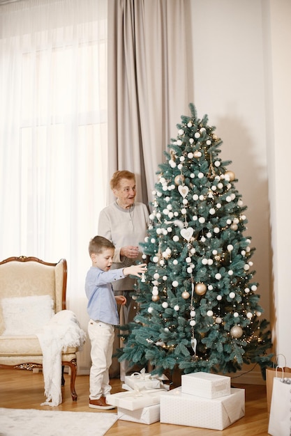 Babcia z wnukiem w Boże Narodzenie. Stara kobieta i mały chłopiec stojący i dekorowanie choinki. Kobieta i chłopiec ubrany w beżowe i niebieskie ubrania.