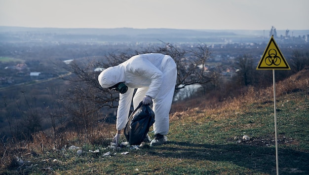 Zdjęcie badacz w kombinezonie ochronnym zbiera plastikowe śmieci do czarnego worka na śmieci na zewnątrz w słoneczny dzień