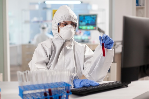 Badacz w kombinezonie trzymający probówki z próbkami krwi pisanymi na komputerze
