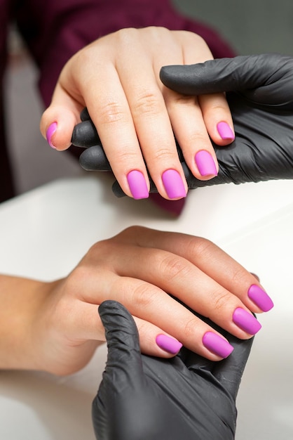 Badanie paznokci wypielęgnowanych. Ręce mistrza manicure w czarnych rękawiczkach, badając kobiece różowe paznokcie w salonie manicure.