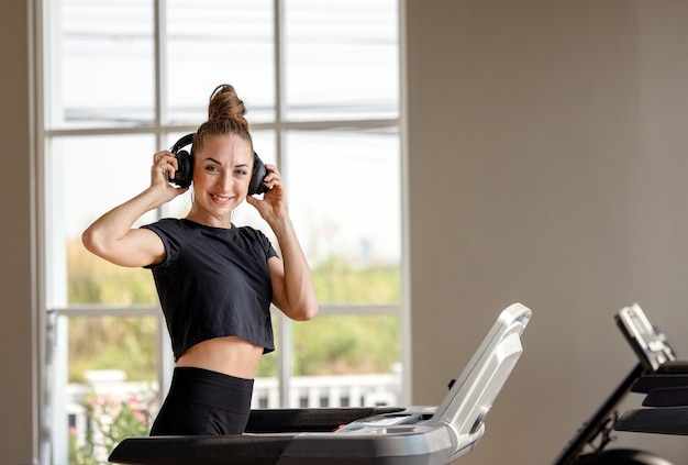 Bądź szybszy! Widok z boku młodej i wesołej kobiety w odzieży sportowej biega na bieżni w siłowni i słuchania muzyki. Trening cardio.