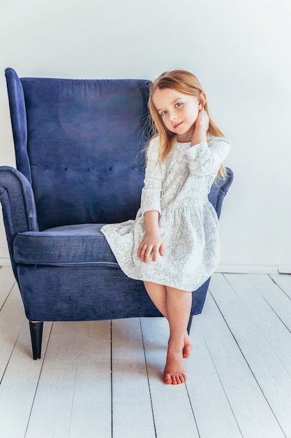 Bądź w domu Bądź bezpieczny. Słodka mała dziewczynka w białej sukni siedzi na nowoczesnym wygodnym niebieskim krześle relaksuje się w białym jasnym salonie w domu w pomieszczeniu. Dzieci w wieku szkolnym młodzież relaks koncepcja.