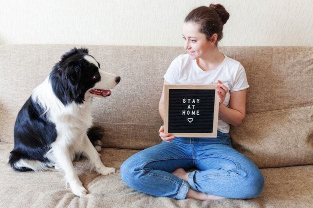 Bądź W Domu Bądź Bezpieczny. Uśmiechnięta Młoda Kobieta Bawić Się Z ślicznym Szczeniaka Psa Border Collie Na Kanapie W Domu Indoors. Dziewczyna Z Napisem Na Tablicy Pobyt W Domu. Koncepcja Kwarantanny życia Zwierząt Domowych Opieki.