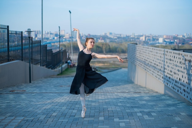 Balerina w tutu pozuje obok ogrodzenia Piękna młoda kobieta w czarnej sukience i pointe tańczy na tle miasta Portret wspaniałej baletnicy wykonującej taniec na świeżym powietrzu