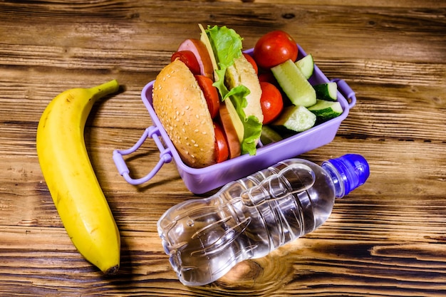 Bananowa butelka wody i pudełko na lunch z ogórkami hamburgerowymi i pomidorami na drewnianym stole