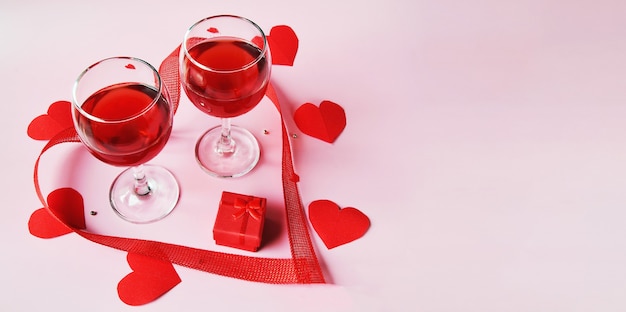 Baner na jasnej powierzchni dwie szklanki czerwonego wina, czerwone pudełko z czerwonymi sercami i wstążką
