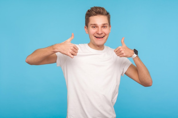 Zdjęcie bardzo dobrze. portret szczęśliwego entuzjastycznego mężczyzny w dorywczo białej koszulce pokazującej kciuki do góry i uśmiechający się przyjaźnie, jest zachwycony zadowolony z wyniku. studio strzał na białym tle na niebieskim tle