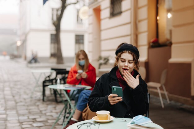 Bardzo młoda brunetka w stylowy ciepły strój, siedząc na tarasie kawiarni na świeżym powietrzu i uśmiechając się, patrząc na ekran smartfona. Maska medyczna i środek odkażający leżący na stole.